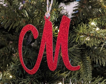 May include: Red glitter letter M Christmas ornament hanging on a green Christmas tree.
