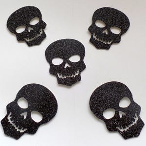 May include: Five black glitter skull shapes, each with a different design, on a white background.