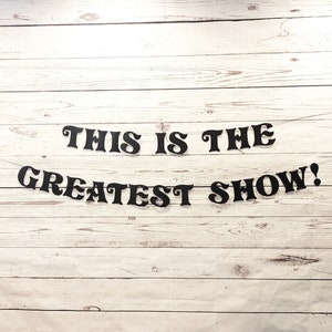 May include: Black letters spelling out "This is the Greatest Show!" on a white wood background.