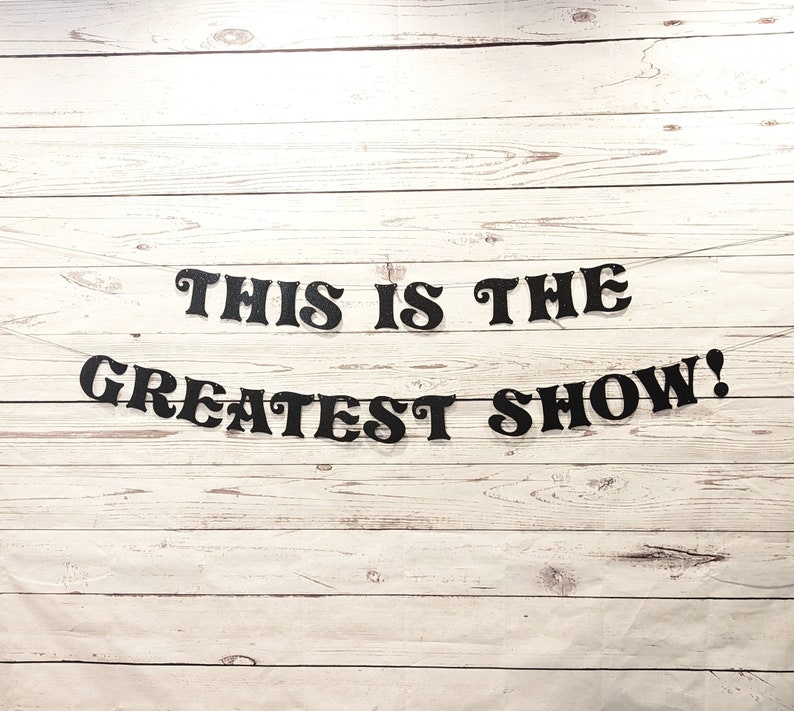 May include: Black letters spelling out "This is the Greatest Show!" on a white wood background.
