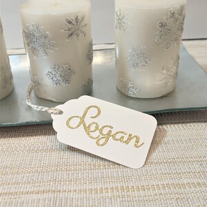 May include: Three white pillar candles with silver snowflake designs on a glass tray. A white gift tag with the name "Logan" written in gold glitter.