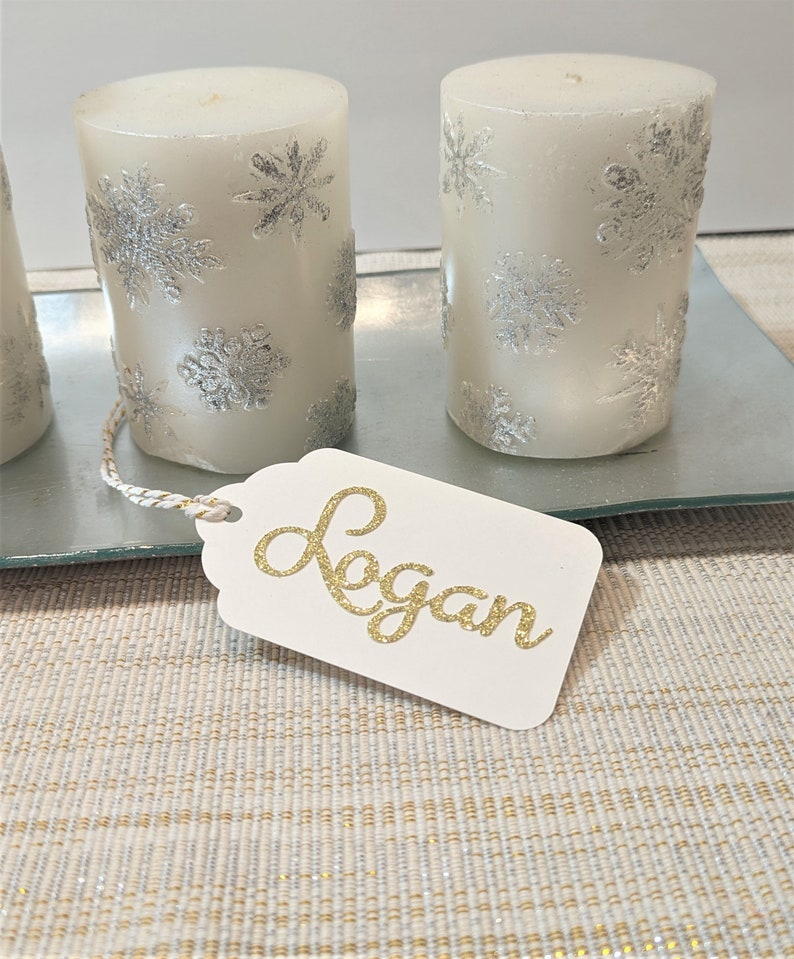 May include: Three white pillar candles with silver snowflake designs on a glass tray. A white gift tag with the name "Logan" written in gold glitter.