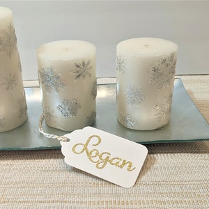 May include: Three white pillar candles with silver snowflake designs on a glass tray. A white gift tag with the name "Logan" written in gold glitter is on the tray.