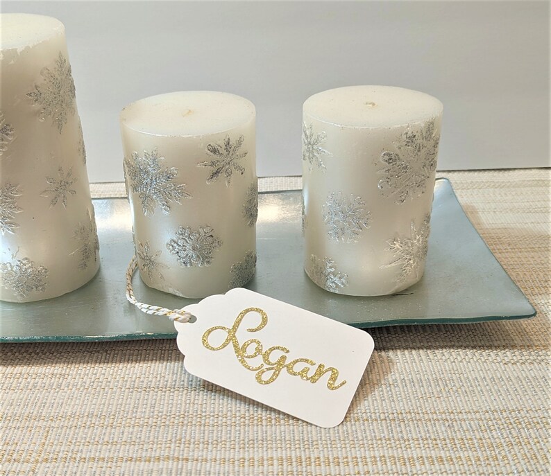 May include: Three white pillar candles with silver snowflake designs on a glass tray. A white gift tag with the name "Logan" written in gold glitter is on the tray.