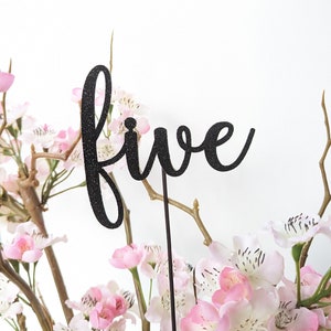 May include: Black glitter "five" table number with a black stick, placed in a bouquet of pink and white flowers.