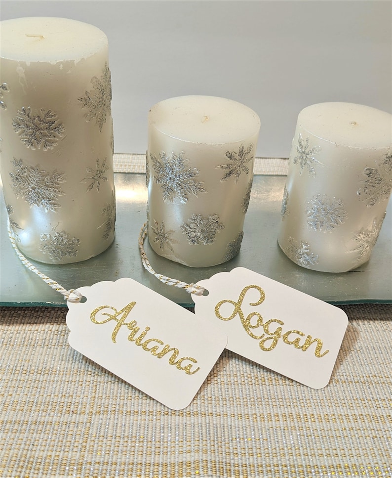May include: Two white candles with silver snowflake designs and gold glitter names "Ariana" and "Logan" on white gift tags.
