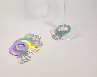 May include: Holographic diamond ring-shaped wine glass charms. The charms are iridescent, reflecting a spectrum of colors. One charm is attached to a wine glass, while several others are grouped together. The design features a diamond shape.