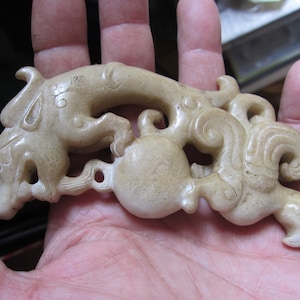 May include: A carved jade pendant depicting a dragon with intricate details. The dragon is curled around a sphere, and the pendant has a hole for hanging.