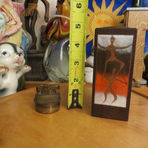 May include: A wooden box with a brown and orange abstract design of a person standing on another person. The box is 5 inches tall.