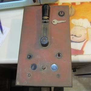 May include: A brass switch panel with a black lever and multiple round buttons. The panel has a red-brown patina and is mounted on a white surface.