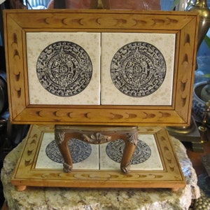 May include: A wooden frame with two ceramic tiles depicting the Aztec calendar, known as the Sun Stone. The tiles are set on a wooden stand with carved details.