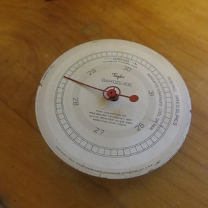 May include: A white circular barometer with a red needle and a red pointer. The barometer is labeled "Tayler BAROGUIDE" and has a scale from 26 to 31. The text "RISING PRESSURES" is printed on the right side of the barometer. The text "INDICATES CONTINUED COOL OR FAIR" is printed on the left side of the barometer.