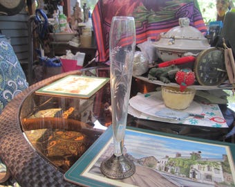 May include: A clear glass vase with etched floral designs. The vase has a slender, elongated shape and a silver-colored base. The vase is displayed on a glass-topped table, with other items in the background.