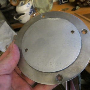 May include: A silver metal round plate with three holes around the edge. The plate is slightly raised in the center.
