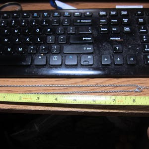 Pu&ograve; includere: Una collana argentata con un ciondolo decorativo e una catena a scatola. La collana &egrave; misurata con un metro a nastro giallo, che mostra una lunghezza di circa 23 cm.