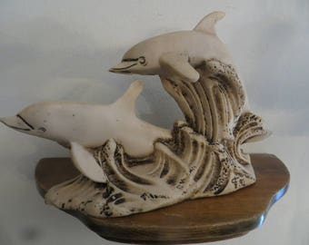 May include: A white ceramic sculpture of two dolphins leaping out of the water. The dolphins are in a wave-like formation, with the larger dolphin in front and the smaller dolphin behind. The sculpture is on a wooden base.