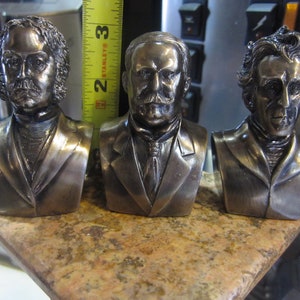 May include: Three silver-toned metal busts of men with different hairstyles and facial features. The busts are displayed on a brown surface.
