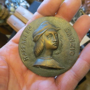May include: A bronze medal with a portrait of Raphael Sanzio, an Italian Renaissance painter. The medal is round and has a raised rim. The portrait is in profile and shows Raphael with a cap and long hair. The inscription "RAFFAELLO SANZIO" is around the edge of the medal.