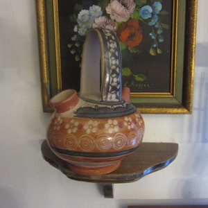 May include: A hand-painted ceramic pitcher with a brown, orange, white, and blue design. The pitcher has a wide, round body and a tall, curved handle. It is sitting on a wooden shelf.