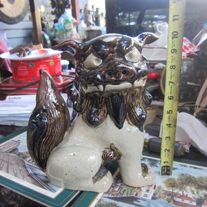May include: A ceramic guardian lion statue, approximately 10 inches tall, with a cream-colored body and detailed brown and black accents. The statue has a fierce expression and a detailed tail.