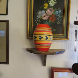 May include: A wooden vase with red, yellow, and black stripes and blue polka dots sits on a wooden shelf. The vase is in front of a framed painting of flowers.