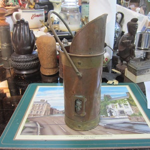 May include: A copper and brass vintage  bucket with a handle. The bucket has a brass band around the top and bottom. The bucket is sitting on a green and white placemat with a picture of a street scene.
