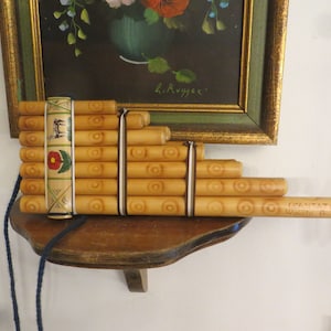 May include: A wooden panpipe with a decorative band featuring a red flower and geometric designs. The panpipe is made of multiple bamboo tubes tied together with brown string. The text "GRAN TAT Walde R." is engraved on the longest tube.