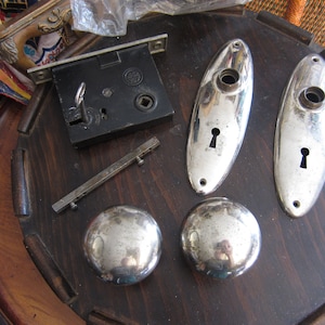 May include: A set of antique door hardware in silver finish. The set includes two oval door plates with keyholes, two round door knobs, a door lock with a key, and a metal rod.