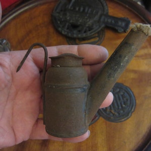 May include: A small, rusty, metal oil lamp with a long spout and a handle. The lamp is held in a hand.