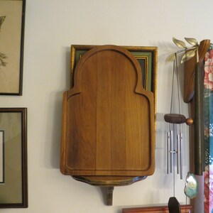 May include: A wooden cutting board with an arched top and a shelf underneath. The board is mounted on a wall and is surrounded by other decor, including a framed picture and a wind chime.