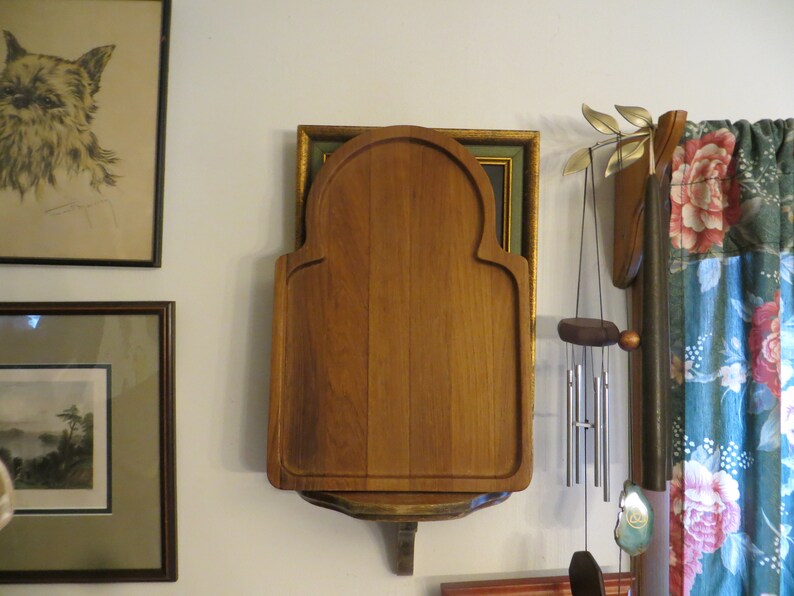 May include: A wooden cutting board with an arched top and a shelf underneath. The board is mounted on a wall and is surrounded by other decor, including a framed picture and a wind chime.