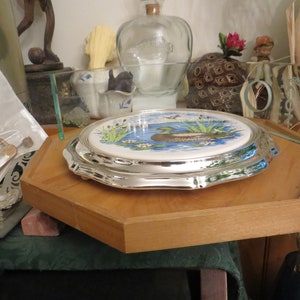 Puede incluir: Un soporte para tartas de plata con una parte superior de cer&aacute;mica blanca que presenta una imagen pintada de un pato en un estanque con juncos verdes y flores blancas.