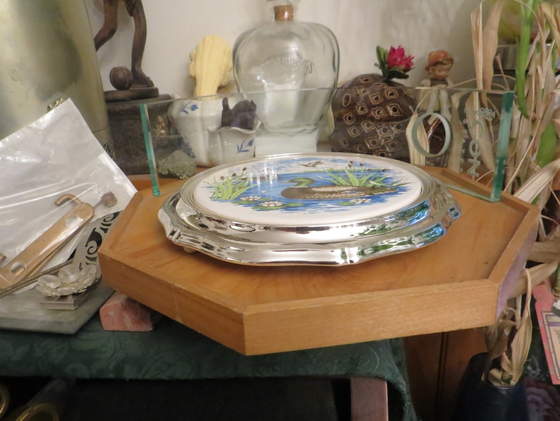 Puede incluir: Un soporte para tartas de plata con una parte superior de cer&aacute;mica blanca que presenta una imagen pintada de un pato en un estanque con juncos verdes y flores blancas.