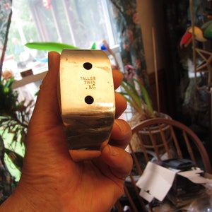 May include: A silver-colored metal cuff bracelet with a rectangular design. The bracelet has two circular holes and the text "TALLER TINTA .EC" is stamped on the surface. The bracelet is held in a hand.