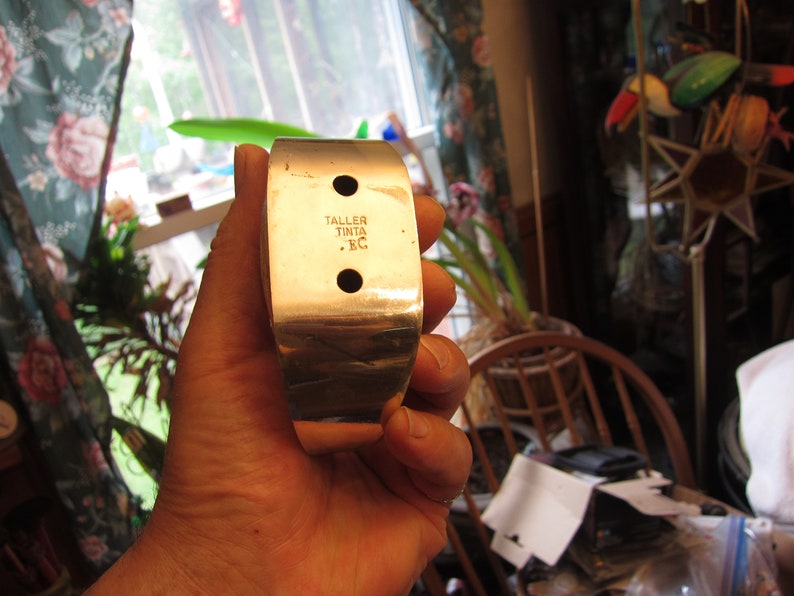 May include: A silver-colored metal cuff bracelet with a rectangular design. The bracelet has two circular holes and the text "TALLER TINTA .EC" is stamped on the surface. The bracelet is held in a hand.