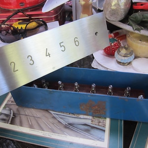 May include: A brushed metal plate with the numbers 1 through 6 etched on it. The plate is held above a blue rectangular box containing several metal components. The background includes various items, suggesting a workshop or storage area.