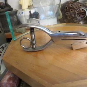 May include: A silver metal punch tool with a spring and a ring handle. The tool is designed to punch holes in paper or other materials.