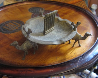 May include: An antique brass ashtray with a central architectural element and three camel figurines. The ashtray has an ornate, etched design and sits on a wooden tray with a dark finish.