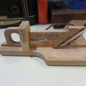 May include: A wooden cheese slicer with a stainless steel blade. The slicer has a handle with a circular opening and a base with a curved edge. The text "So Apéro!" is visible on the wooden surface. A label reads "The French Guillotine".