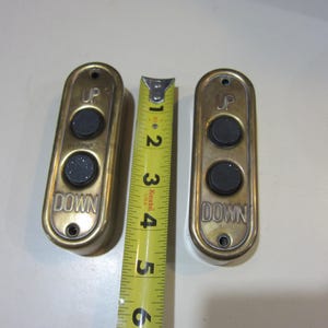 May include: Two vintage brass elevator call buttons with "UP" and "DOWN" text, each with two black buttons. A yellow measuring tape shows the buttons are approximately 5 inches long.