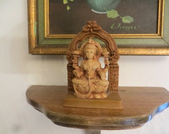 May include: A wooden figurine of a seated goddess with a crown and ornate jewelry. The figurine is on a wooden shelf with a framed painting behind it. The painting is signed "L. Ruyter".