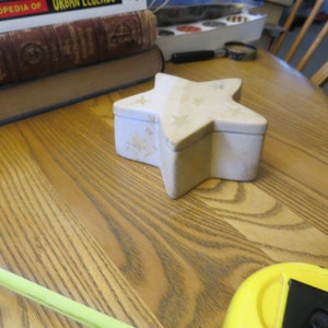 May include: A white star-shaped trinket box with a white star design on the lid. The box is sitting on a wooden surface.