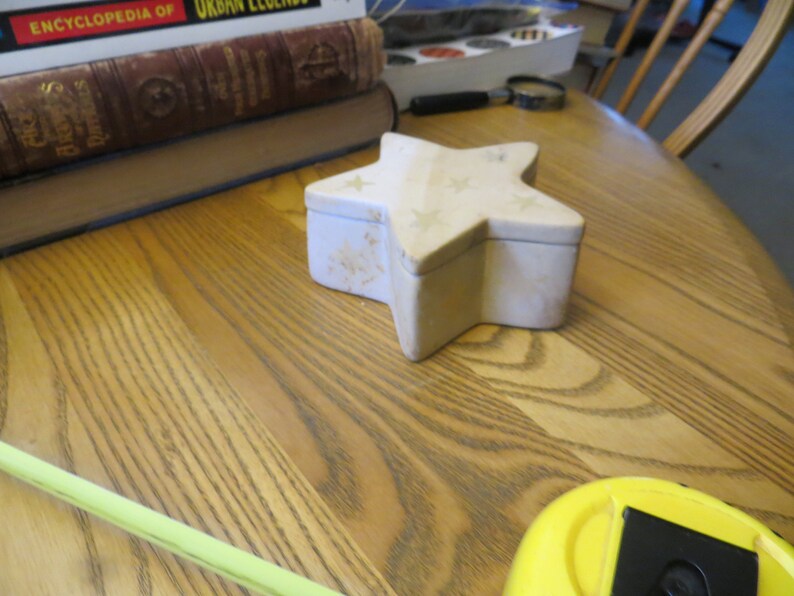 May include: A white star-shaped trinket box with a white star design on the lid. The box is sitting on a wooden surface.