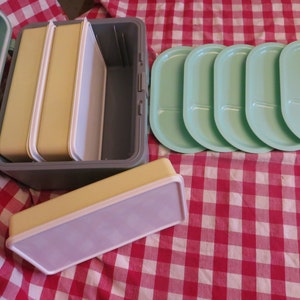 May include: A light blue lunchbox with three yellow and white containers inside. Four light green divided plates are next to the lunchbox.