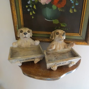 May include: Two ceramic dog figurines holding small wooden trays. The dogs are beige with black eyes and noses. The trays have the words "Milk Money" and "For Our Kids" written on them.
