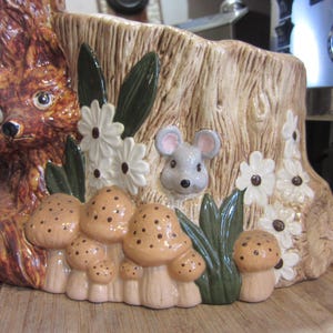 May include: A ceramic planter shaped like a tree stump with a squirrel, a mouse, mushrooms, and flowers. The planter is painted in shades of brown, green, white, and gray.