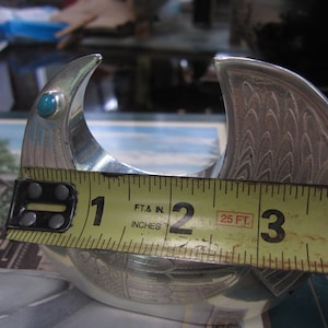 May include: A silver-colored metal object with a curved design, featuring a turquoise stone. The object is approximately 3 inches wide. The design includes etched patterns resembling feathers. The object is likely a decorative item.