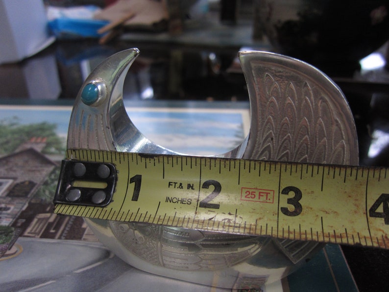 May include: A silver-colored metal object with a curved design, featuring a turquoise stone. The object is approximately 3 inches wide. The design includes etched patterns resembling feathers. The object is likely a decorative item.
