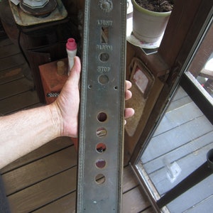 May include: A vintage brass elevator control panel with buttons labeled "Light", "Alarm", and "Stop". The panel has a decorative border and is held in a hand.