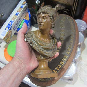 May include: Bronze bust of a woman wearing a helmet, mounted on a wooden plaque. The bust has a detailed face and draped clothing. The plaque has gold lettering that reads "THE EIGHT ELEVATOR".